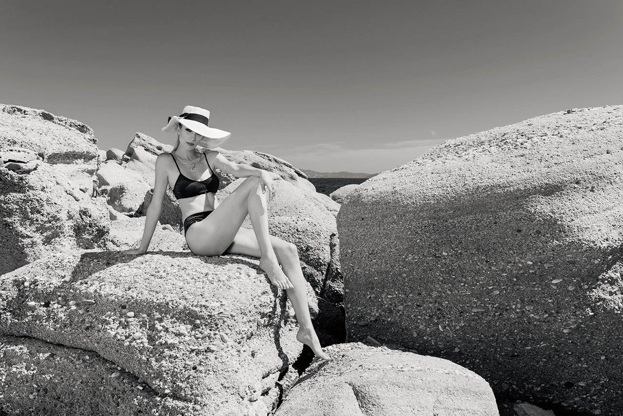 Fotografia in bianco e nero di una modella con costume a due pezzi, cappello, collana e bracciale sul bicipite, seduta sugli scogli con il mare sullo sfondo.